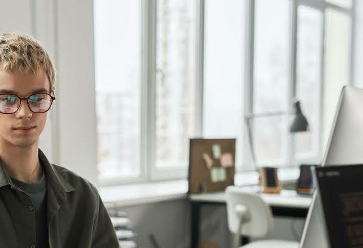 Mann med briller sitter ved skrivepulten og jobber foran laptop og skjerm
